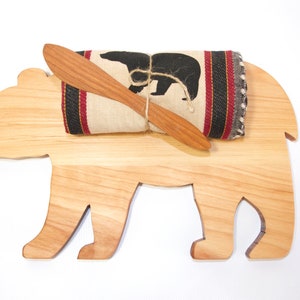 May include: Wooden bear-shaped cutting board with a rolled up tea towel and a wooden butter knife on top. The tea towel has a black bear silhouette and red and black stripes.