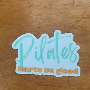 white and teal Pilates sticker that says Pilates hurts so good in a script font 3 x 3 inches