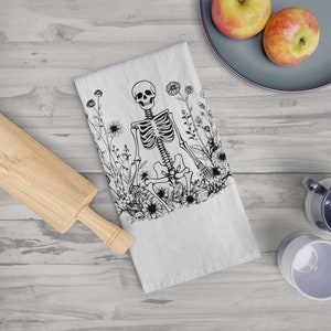 May include: A black and white kitchen towel with a skeleton sitting in a field of flowers. The skeleton is sitting with its legs crossed and its arms resting on its knees. The flowers are in full bloom and are surrounding the skeleton.