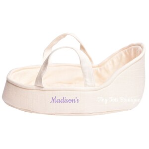 May include: A cream-colored doll bassinet with a white and cream striped lining. The bassinet has two handles and is embroidered with the text "Madison's Tiny Tote Boutique".