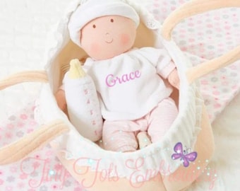 Puede incluir: Una muñeca de peluche con gorro blanco y camiseta blanca con el nombre Grace en letra cursiva rosa, vistiendo pantalones rosa claro. La muñeca sostiene un biberón de juguete blanco con marcas rosas. Está dentro de un moisés de tela color crema.