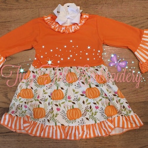 Puede incluir: Vestido naranja de manga larga con lazo blanco. La parte superior es naranja con cuello con volantes y detalles de estrellas. La falda presenta un estampado de calabaza y flores con volantes a rayas naranjas y blancas. Las mangas tienen volantes a rayas y lunares.