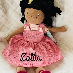 May include: A plush doll with dark skin, black curly hair, and a gold bow. The doll wears a pink dress with a red plaid pattern and a cat face design. The name "Lolita" is embroidered on the dress.