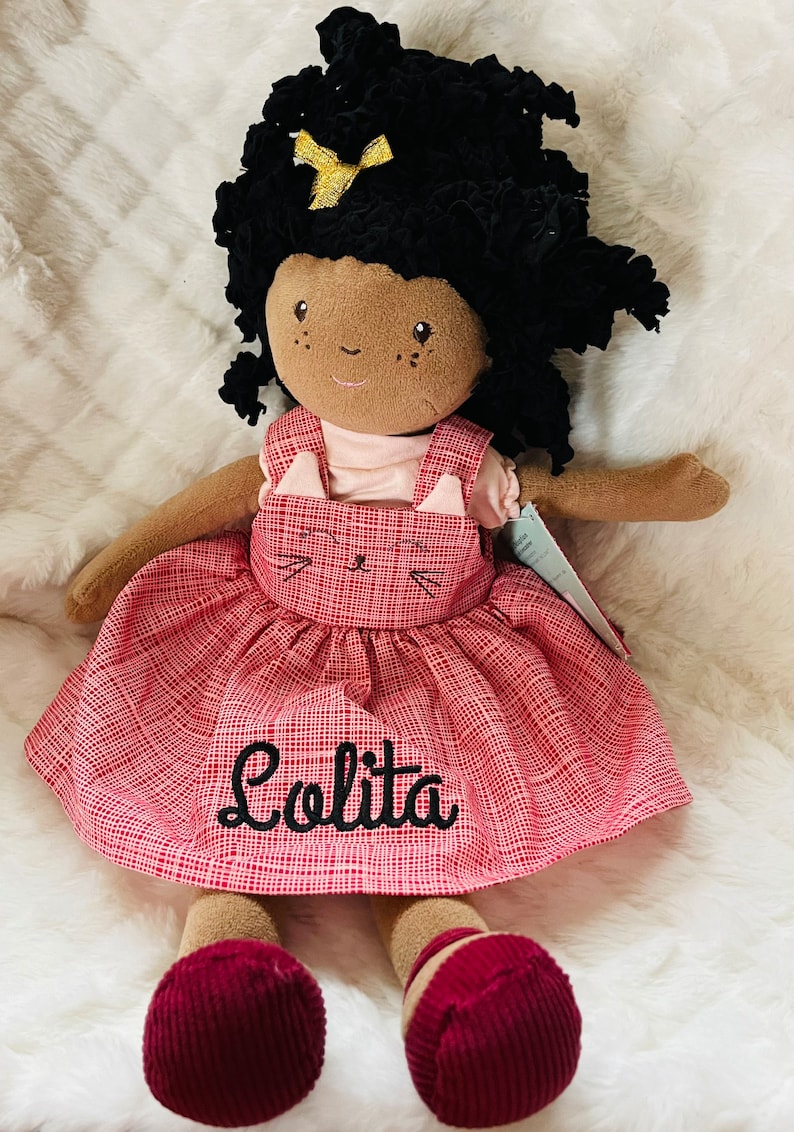 May include: A plush doll with dark skin, black curly hair, and a gold bow. The doll wears a pink dress with a red plaid pattern and a cat face design. The name "Lolita" is embroidered on the dress.