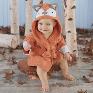 May include: A baby wearing an orange fox-themed hooded bathrobe, with a white and gray fox face on the hood. The baby is sitting on a wooden stand, with a background of birch trees and fallen leaves.