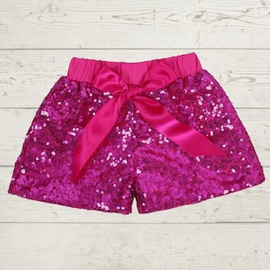 May include: Hot pink sequin shorts with a matching bow. The shorts have an elastic waistband and are covered in shiny sequins. The bow is made of a satin-like material and is tied in the front. The shorts are a vibrant, eye-catching piece.