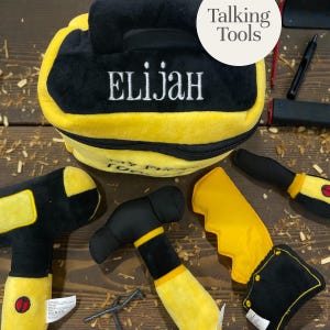 Puede incluir: Un juego de herramientas de juguete de felpa negra y amarilla, que incluye una bolsa bordada con el nombre "ELIJAH". El juego incluye herramientas de juguete como un martillo, una sierra y un destornillador. La bolsa tiene el texto "Talking Tools".