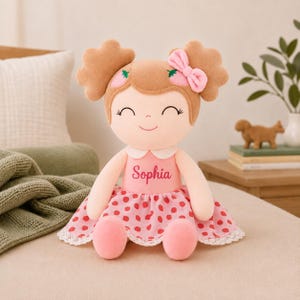 May include: A plush doll with light brown hair styled in two buns, featuring a pink bow and strawberry accents. The doll wears a pink dress with a red strawberry pattern, a white collar, and lace trim. The name Sophia is embroidered in pink on the chest.