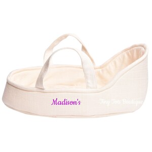 May include: A cream-colored doll bassinet with two handles and a striped lining. The bassinet has the text "Madison's" and "Tiny Tote Boutique" printed on it.