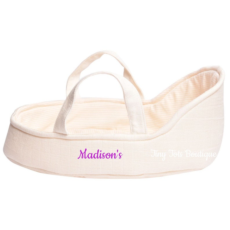 May include: A cream-colored doll bassinet with two handles and a striped lining. The bassinet has the text "Madison's" and "Tiny Tote Boutique" printed on it.