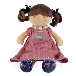 Könnte beinhalten: Eine Stoffpuppe mit braunen Haaren in Zöpfen, die mit dunkelblauen Bändern gebunden sind. Sie trägt ein altrosa Kleid, auf dem der Name Mandy in Türkis gestickt ist. Die Puppe hat eine Perlenkette in Mint und Rosa sowie dunkelblaue Stoffschuhe.