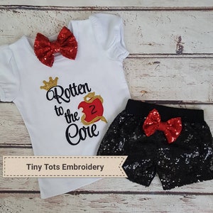 Puede incluir: Un conjunto de camiseta blanca y shorts de lentejuelas negras con lazos rojos de lentejuelas. La camiseta presenta el texto "Rotten to the Core" con un gráfico de manzana y una corona dorada. Los shorts tienen una cintura elástica negra.