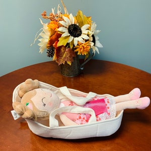 May include: A white fabric doll carrier with a plush doll wearing a pink and white dress with red polka dots. The doll has brown hair and closed eyes. The doll carrier has two straps and is sitting on a wooden table.
