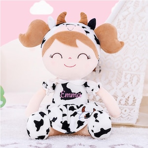 May include: A plush doll with a cow-themed outfit. The doll has brown hair, a smiling face, and wears a dress and hat with black and white cow spots. The name "Emma" is printed on the dress.