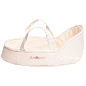 May include: A cream-colored doll bassinet with a white and cream striped lining. The bassinet has two handles and is embroidered with the text "Madison's Tiny Tote Boutique".
