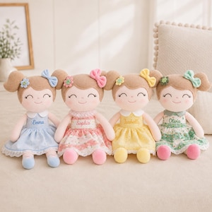 May include: Four plush dolls with brown hair in pigtails, each wearing a dress with a lace hem and a matching hair bow. The dolls are embroidered with the names Emma, Sophia, Isabella, and Olivia on their bodices, featuring floral patterns.
