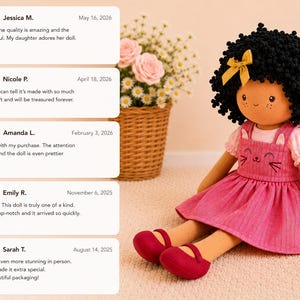May include: A handmade doll with black curly hair, a yellow bow, and a pink dress with a cat face design. The doll is wearing red shoes and is sitting on a light-colored surface. Reviews are visible in the background.