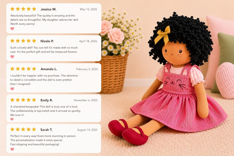 May include: A handmade doll with black curly hair, a yellow bow, and a pink dress with a cat face design. The doll is wearing red shoes and is sitting on a light-colored surface. Reviews are visible in the background.