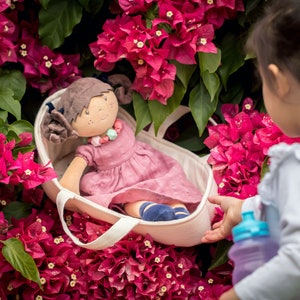 May include: A little girl is holding a white canvas tote bag with a doll inside. The doll is wearing a pink dress and has brown hair. The bag is surrounded by pink flowers.