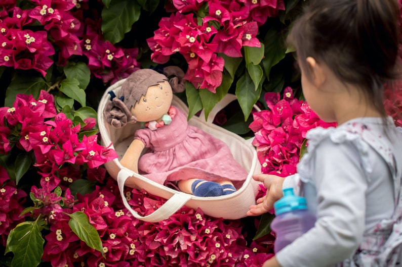 May include: A little girl is holding a white canvas tote bag with a doll inside. The doll is wearing a pink dress and has brown hair. The bag is surrounded by pink flowers.
