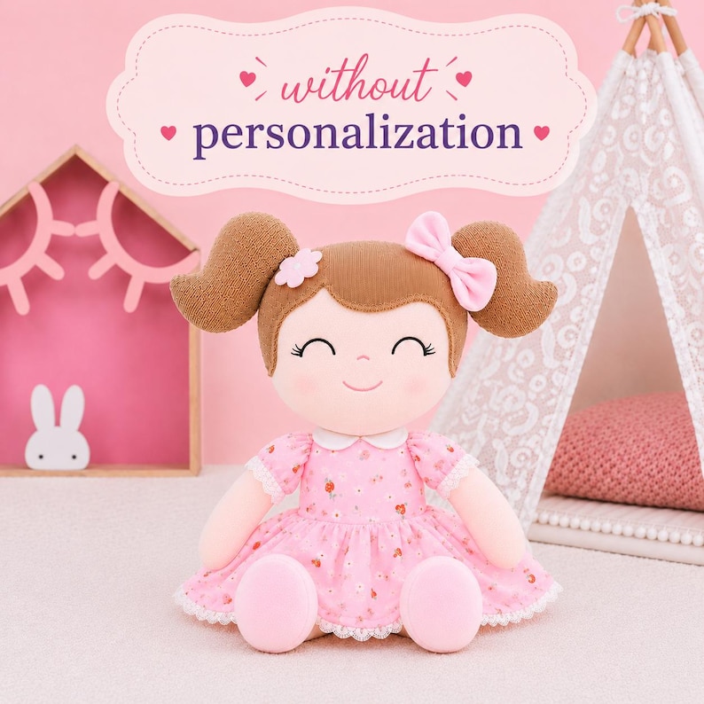 May include: A plush doll with brown hair styled in pigtails, wearing a pink floral dress with white lace trim and a small pink bow. The doll has a smiling face and is positioned below text that reads without personalization in a decorative frame.