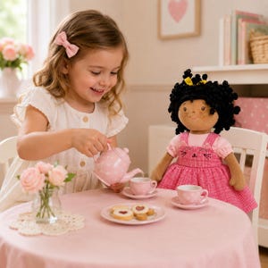 May include: A young girl pours from a pink polka-dot teapot into a matching teacup. A doll with black curly hair and a pink dress sits at the table. The table is set with a plate of cookies and a vase of pink roses, creating a charming tea party scene.