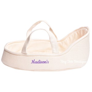 May include: A cream-colored fabric doll bassinet with a white and cream striped lining. The bassinet has two fabric handles and is embroidered with the text "Madison's" and "Tiny Tote Boutique".