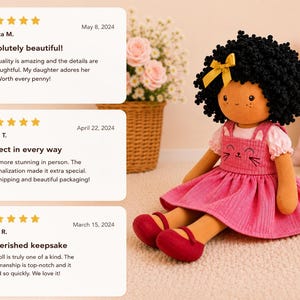 May include: A handmade doll with a black curly hairstyle and a yellow bow. The doll wears a pink dress with a cat face design and red shoes. The doll is sitting on a light-colored surface.