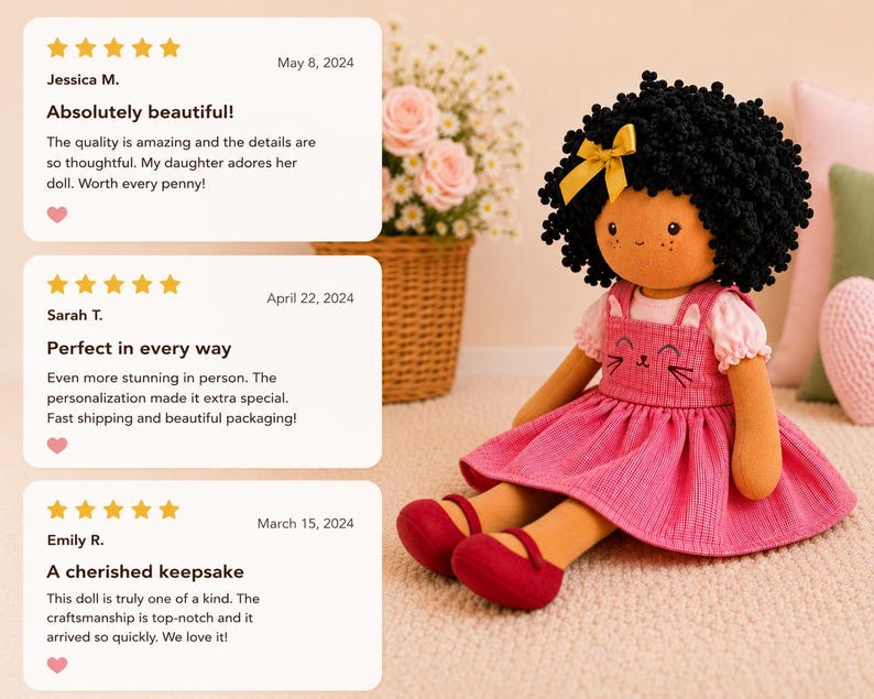 May include: A handmade doll with a black curly hairstyle and a yellow bow. The doll wears a pink dress with a cat face design and red shoes. The doll is sitting on a light-colored surface.