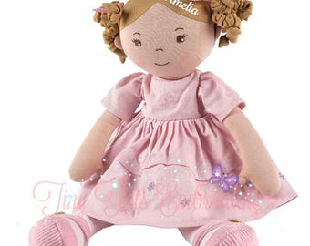 Könnte beinhalten: Eine Stoffpuppe mit hellbraunem Haar, das zu zwei Zöpfen mit rosa Schleifen gebunden ist. Auf dem Kopf ist der Name Amelia eingestickt. Die Puppe trägt ein hellrosa Kleid mit kurzen Ärmeln, einem gestuften Rock und dazu passende rosa Schuhe.