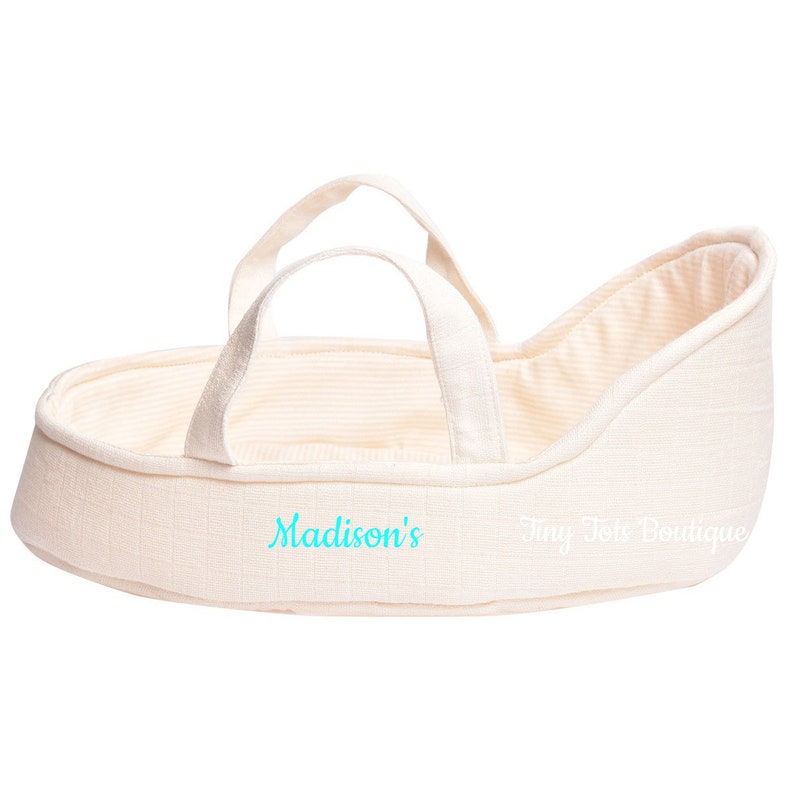 May include: A cream-colored doll bassinet with a white and cream striped lining. The bassinet has two handles and is embroidered with the text "Madison's Tiny Tote Boutique".