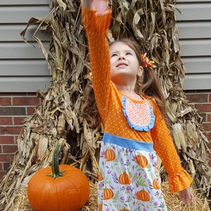 Puede incluir: Un conjunto otoñal con una blusa naranja de lunares, una falda con estampado de calabazas y leggings a rayas. Una gran calabaza naranja sobre una paca de heno. Ideal para Halloween.