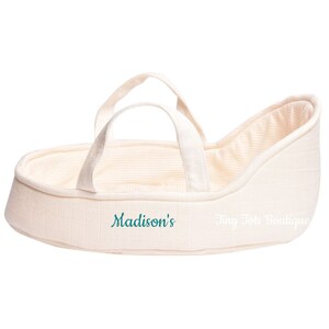 May include: A cream-colored doll bassinet with a white and cream striped lining. The bassinet has two handles and is embroidered with the text "Madison's Tiny Tote Boutique".