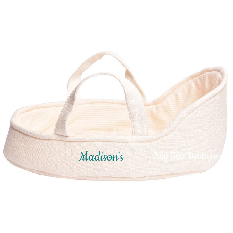 May include: A cream-colored doll bassinet with a white and cream striped lining. The bassinet has two handles and is embroidered with the text "Madison's Tiny Tote Boutique".