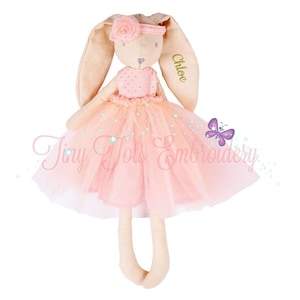 May include: A plush rabbit doll with long ears, wearing a peach-colored dress with a tulle skirt and a matching headband featuring a fabric flower. The name Chloe is embroidered in gold on one ear. The text Tiny Tots Embroidery is visible.