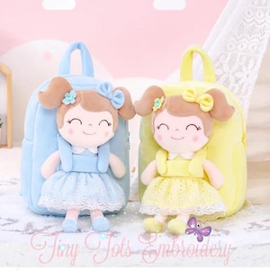 May include: Two plush backpacks, one light blue and one yellow, each featuring a soft doll attached to the front. The dolls have brown hair with bows, smiling faces, and dresses with lace trim. The text Tiny Tots Embroidery is visible below.