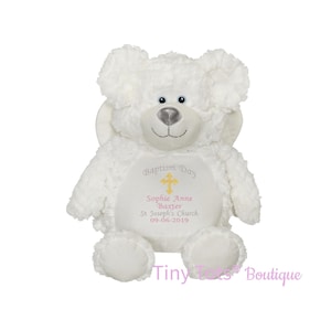 Puede incluir: Un oso de peluche blanco con una cara sonriente y ojos azules. El oso tiene las palabras "Baptism Day" y una cruz bordada en el pecho, junto con un nombre y una fecha. Un juguete suave y tierno.