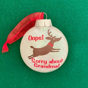 May include: White glitter Christmas ornament with a brown reindeer jumping and the text "Oops! Sorry about Grandma!" in red.