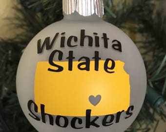 May include: A frosted glass Christmas ornament with the text "Wichita State Shockers" and a yellow outline of the state of Kansas with a small gray heart in the center.