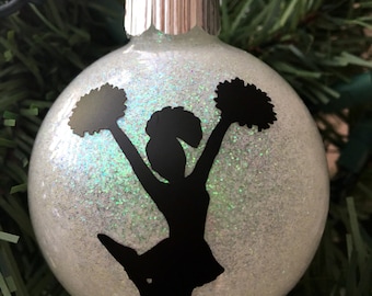May include: A white glass Christmas ornament with a glittery finish. A black silhouette of a cheerleader holding pom-poms is on the ornament.