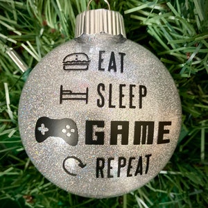 May include: Silver glitter Christmas ornament with black text that reads "Eat Sleep Game Repeat" and icons for a hamburger, bed, and video game controller.