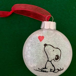 May include: A white glitter Christmas ornament with a black and white illustration of Snoopy looking up at a red heart. The ornament has a red ribbon attached to the top.