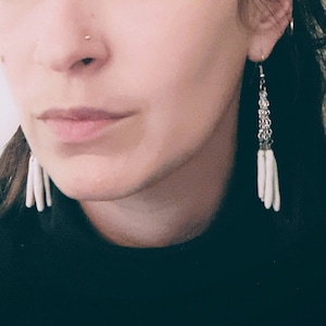 Può includere: Primo piano di orecchini a catena argentata con pendenti bianchi allungati a forma di nappa. Gli orecchini sono indossati da una persona con un top nero.