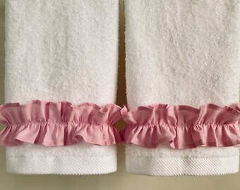 Puede incluir: Dos toallas de baño blancas con ribete de volantes rosa colgadas de un toallero plateado.