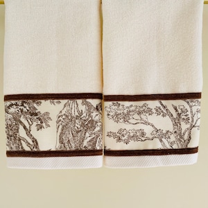 May include: Two white bath towels with brown trim and a toile pattern. The pattern features brown trees and figures on a white background.