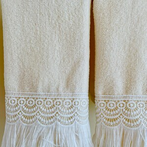 May include: Two cream-colored towels with white lace trim and fringe. The towels are hanging on a towel rack.