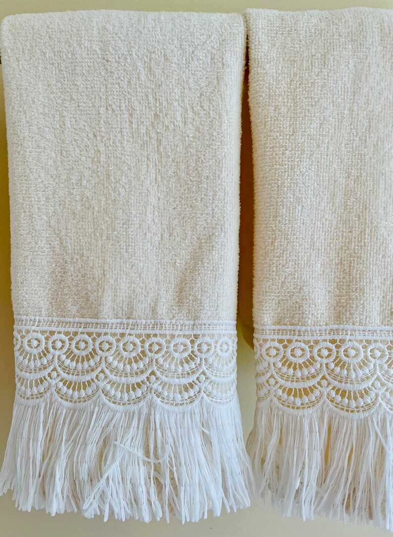 May include: Two cream-colored towels with white lace trim and fringe. The towels are hanging on a towel rack.