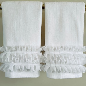 May include: Two white bath towels with a textured surface, hanging on a gold-colored bar. The bottom of each towel is adorned with two rows of ruffled, sheer fabric, adding a decorative touch. The towels are clean and neatly arranged.