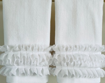 May include: Two white bath towels with a textured surface, hanging on a gold-colored bar. The bottom of each towel is adorned with two rows of ruffled, sheer fabric, adding a decorative touch. The towels are clean and neatly arranged.