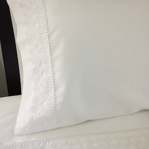 May include: White pillowcase with a white lace trim around the edge. The lace trim is a delicate pattern with a scalloped edge.